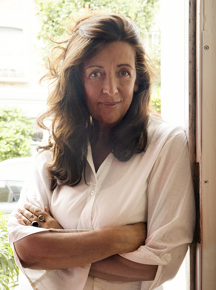 A photograph of Louise leaning against a door frame. Louise wears a white shirt and her hair falls just below shoulder length. She looks directly at the camera with a soft smile.