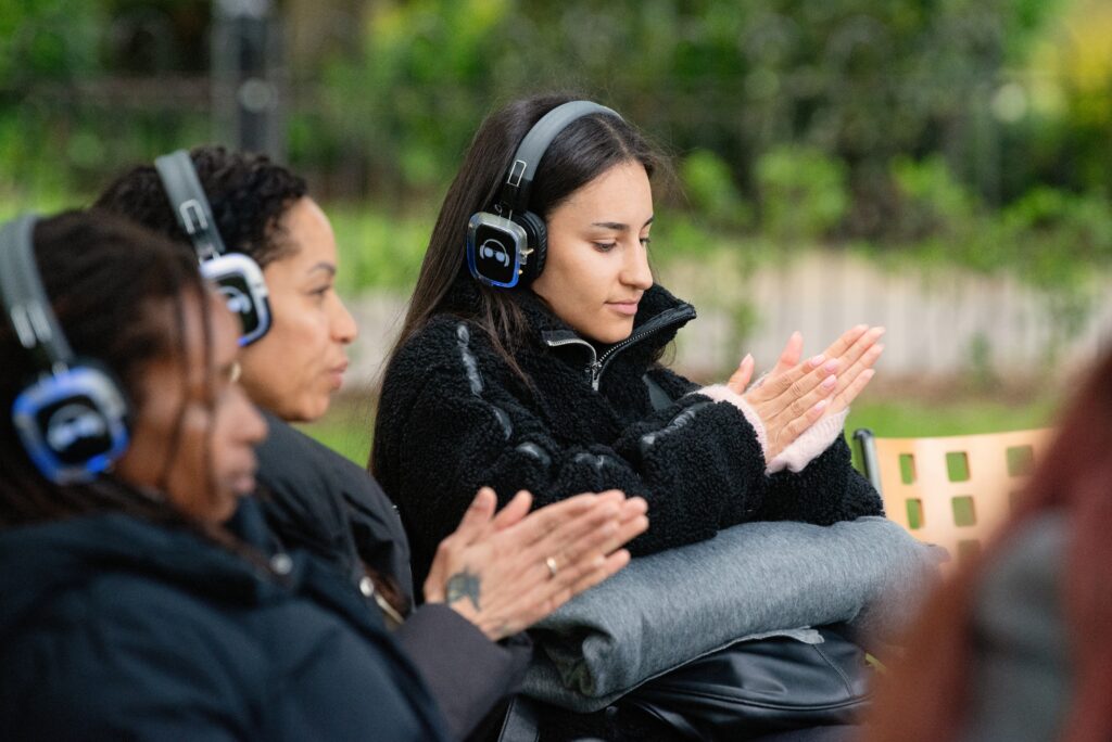 Three people sitting in an outdoor setting. They have headphones on and are all rubbing their hands together.