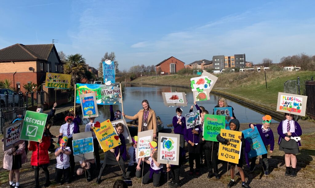 A group of school children stand by a canal and wave placards with slogans about recyling on them