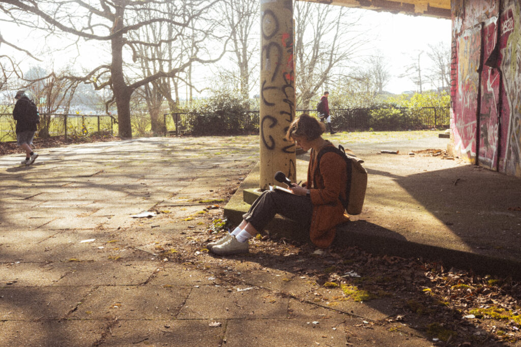 Someone sits holding a microphone in an outdoor location - a building covered in graffiti - bathed in sunlight. There is a park in the distance.