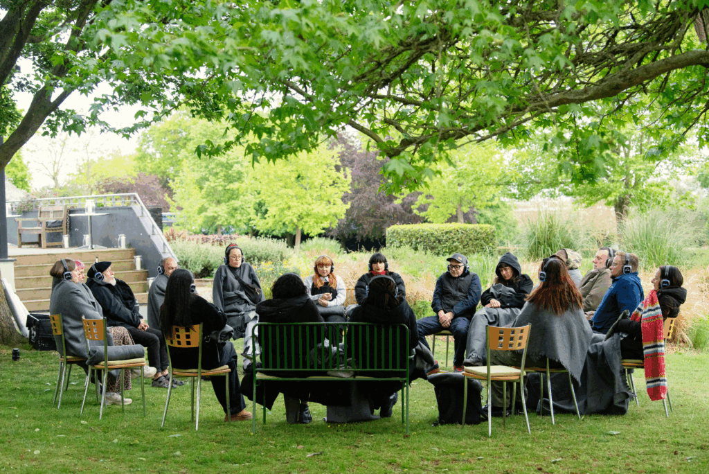 A group of people sit in a circle in a beautiful green outdoor space under a tree. They are wearing headphones and have their eyes closed.