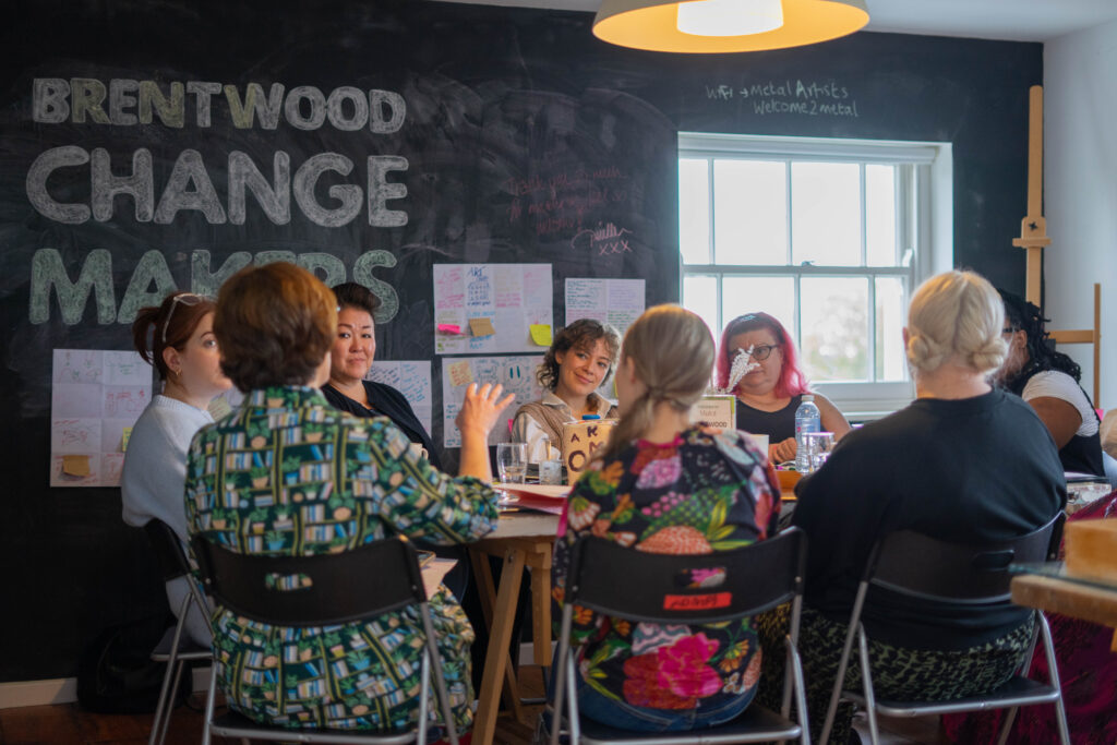 A group of people sit around a table talking and smiling. The words Brentwood Change makers is on the wall behind them