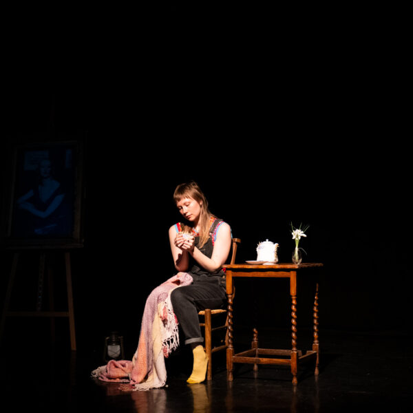 A photo of performer Louise Kim Salter sitting on a chair on stage, with a table besides them, with a blanket draped on their knee.