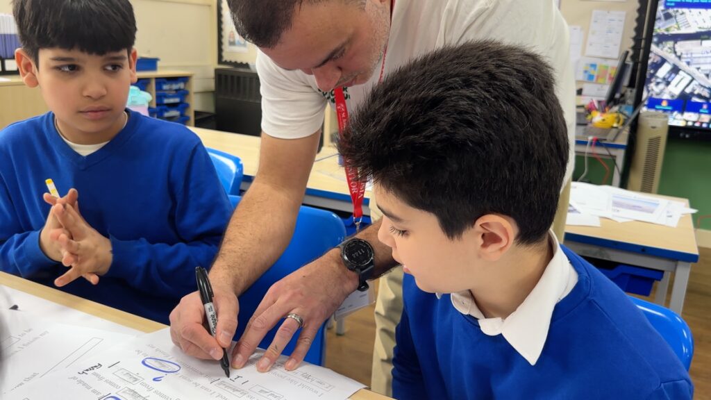 two boys in school uniform are watching a man as he draws a diagram on a large sheet of paper on a desk