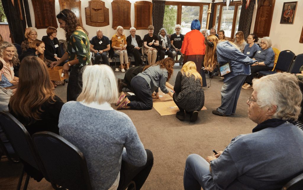 A group of about 25 people sit on chairs in a circle in an indoor space. There are some people kneeling on the floor adding post its to a large sheet of bown paper while other stand beside them peering over to look.
