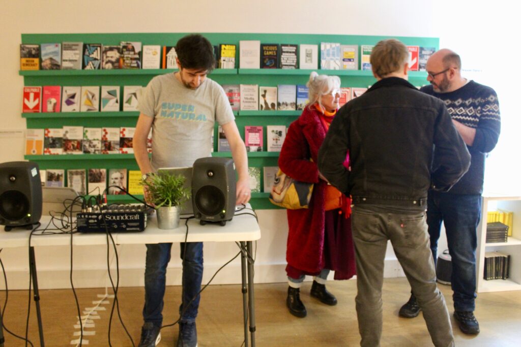 Dreamspace Peterborough - a bright room with colourful green bookshelves covered in a variety of books on cultural themes with the covers facing out. A man stands in front of the shelves playing music through a laptop on a table with speakers eaither side of him. To his right a group of poeple are standing nad chatting together.